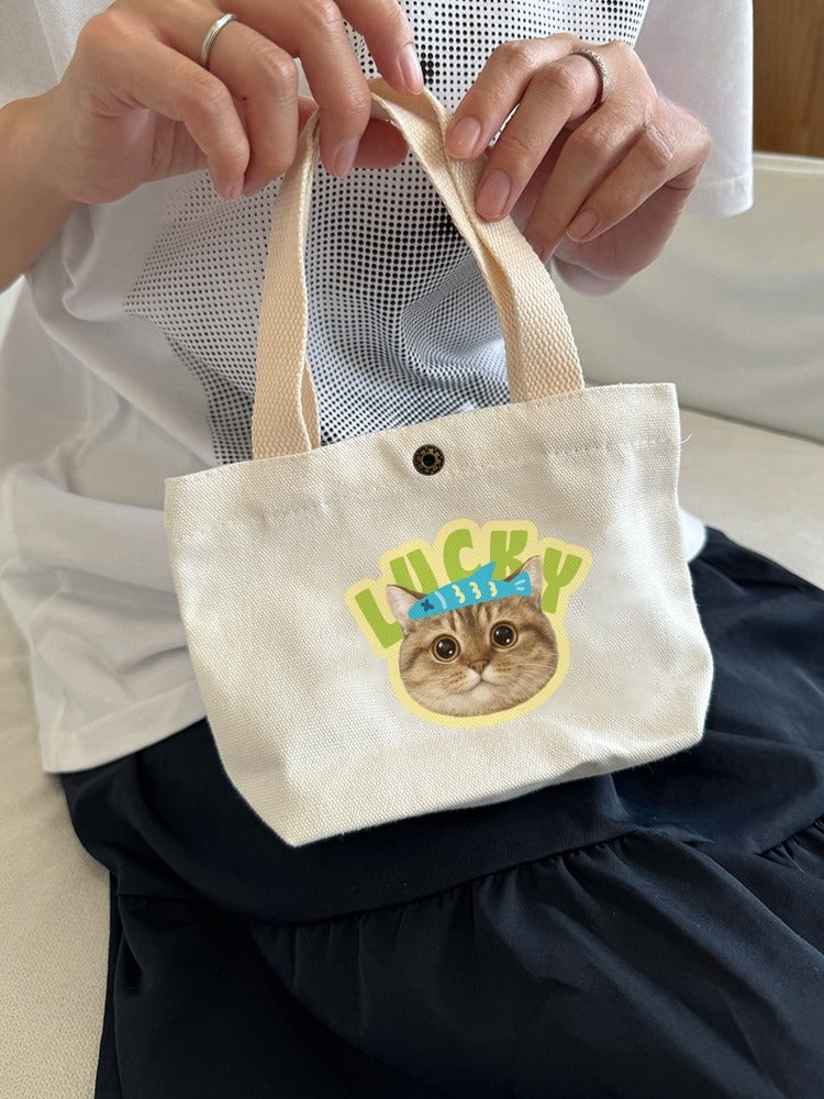 White tote bag with a cat graphic and 'Lucky' text held by a person.